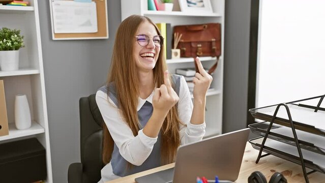 Blonde young businesswoman shows off 'fuck you' sign boldly at office, exhibits bad attitude with middle finger, provokes with rude gesture, excitedly expressing workplace rudeness indoors.