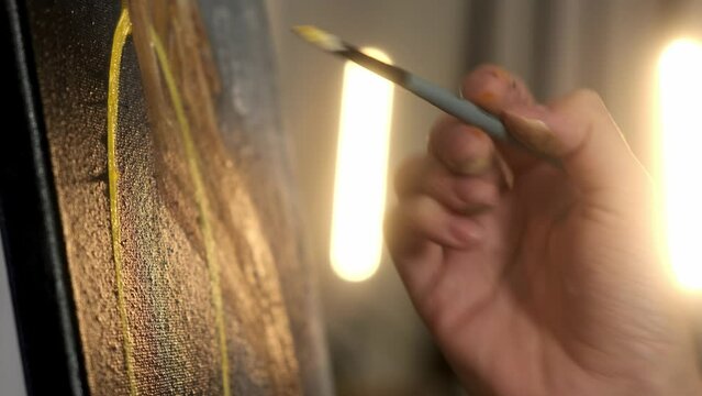 Female artist draws drawings, creating new works of art on black canvas, enjoying the process of painting. Narrow brush for applying yellow gold oil paint. Choosing materials color palette close-up