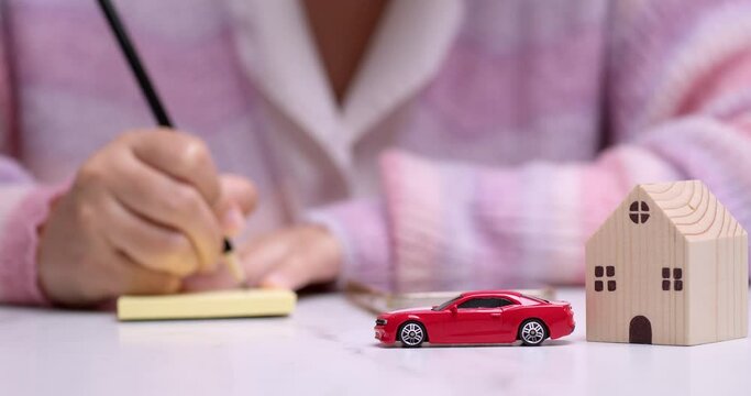 Close-up of a hand holding a pencil and writing something on a yellow paper, behind a red toy car that signifies car loan financing.