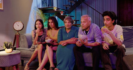 Indian Asian family friends aged mom dad man sitting sofa watch horror thriller TV scared old elder...