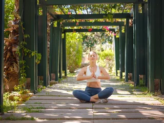 Foto op Plexiglas Happy adorable athletic woman performing yoga sitting in lotus position outdoors against background of green city park, sporty female during meditation practice. sukhasana, healthy lifestyle concept. © YURII Seleznov