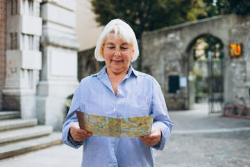 Mature blonde woman tourist looking at city map at street. Cheerful 60s woman traveling abroad in...