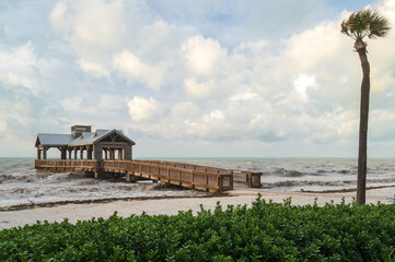 A wooden pier, with a covered portion at the end, reaching out into a choppy, rough sea, with tropical foliage in the foreground