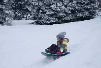 Fototapeta na wymiar Little girl with rainbow jacket sledging down a snowy hill. High quality photo