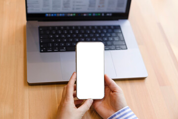 A smartphone in the hands of a woman with a white background, a laptop on the table. Phone with space for copy, work in the office at home or in a cafe, freelancer, hands close-up