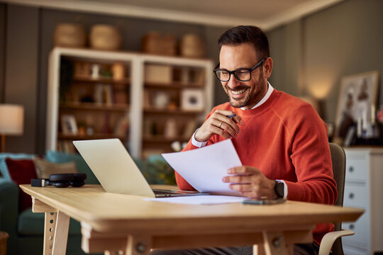 A smiling male remote worker comparing a document in his hand to the one on his laptop.