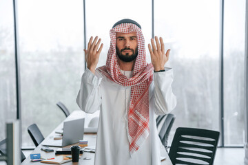 Praying with hands near the face. Successful Muslim businessman in traditional outfit in his office