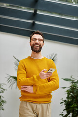 cheerful handsome architect with glasses in yellow turtleneck posing with his phone in his office