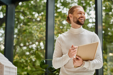 cheerful handsome man in white casual turtleneck with beard posing in his office while working hard