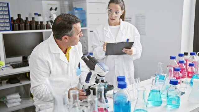 Man and woman scientists high five in lab, writing on clipboard with microscope, celebrating a smiling medical discovery together