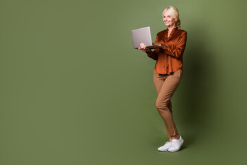 Full length photo of lovely senior lady hold netbook eshopping promo dressed stylish brown silk garment isolated on khaki color background