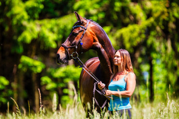 A young woman rider with long brunette hair stands with her horse on a high summer meadow in the...