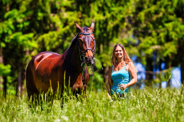 A young woman rider with long brunette hair stands with her horse on a high summer meadow in the...