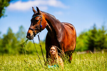 A young woman rider with long brunette hair stands with her horse on a high summer meadow in the...