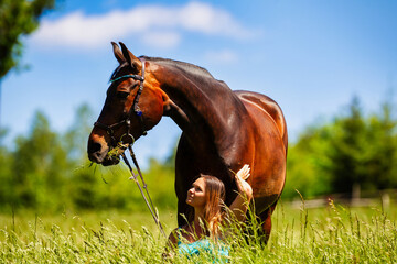 A young woman rider with long brunette hair stands with her horse on a high summer meadow in the bright sunshine.