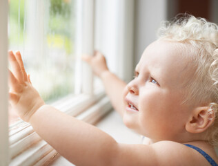 Baby, window and child thinking by glass at home, curious and sad alone. Young kid, blonde toddler...