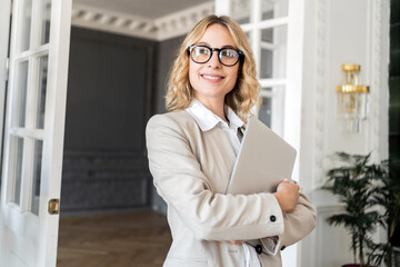 Smiling businesswoman holding a laptop, standing confidently in an elegant interior.