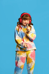 Little happy girl with down syndrome wearing headphones, holing microphone and singing against blue...