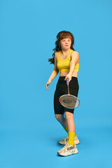 Full-length image of teen girl with down syndrome in sportswear playing badminton, training over blue studio background. Concept of acceptance, care, inclusion, health, diversity, emotions, equality
