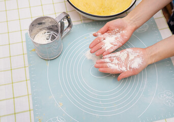 The image shows hands covered in flour over a silicone baking mat. A sifter and mixing bowl are nearby.