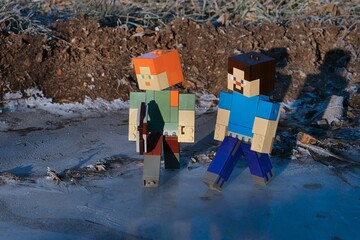 Fototapeta premium LEGO Minecraft main characters Alex (with iron pickaxe) and smiling Steve walking across icy frozen water puddle during cold winter morning.