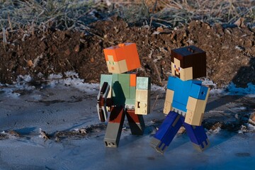 Fototapeta premium LEGO Minecraft main characters Alex (with iron pickaxe) and smiling Steve walking on icy frozen water puddle during cold winter morning.