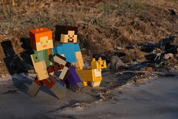 Fototapeta premium LEGO Minecraft figures of Alex with iron pickaxe, smiling Steve and pet friend Ocelot walking across icy surface in winter morning sunshine. 