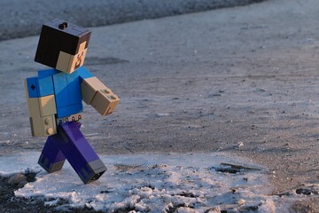 Obraz premium LEGO Minecraft figure of Steve walking along frozen water surface near asphalt road, winter morning sunshine. 