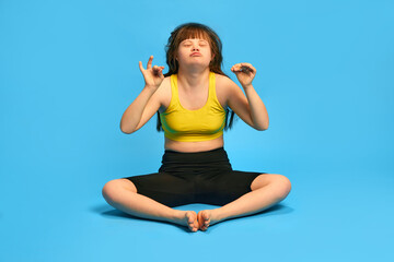 Therapy. Feeling calm and relaxed. Teen girl with down syndrome sitting in yoga pose against blue studio background. Concept of acceptance, care, inclusion, health, diversity, emotions, equality