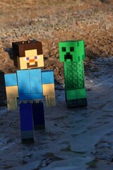 Fototapeta premium LEGO Minecraft figure of Steve chased by green explosive Creeper mob on icy surface of frozen puddle, morning winter sunlight. 