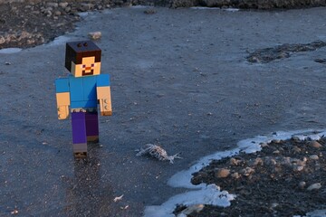 Fototapeta premium LEGO Minecraft figure of Steve walking on ice frosty surface near asphalt road, sunlit by early morning winter sunshine.