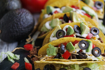 Tacos made of corn tortillas, ground beef, shredded cheddar cheese, black olives, avocados, lettuce, sour cream and tomatoes. Selective focus with blurred foreground and background.