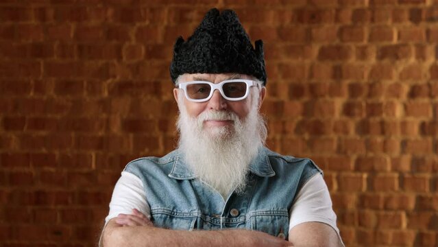 Stylish grandfather in a black woolen hat and white glasses, posing for the camera against a brick wall. He is dressed in a denim vest and a white t-shirt. Camera 8K RAW.