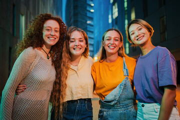 Portrait of a group of young women smiling looking at camera together at night time. Four female friends standing outside with happy expression. Proud feminine ladies staring front at social gathering