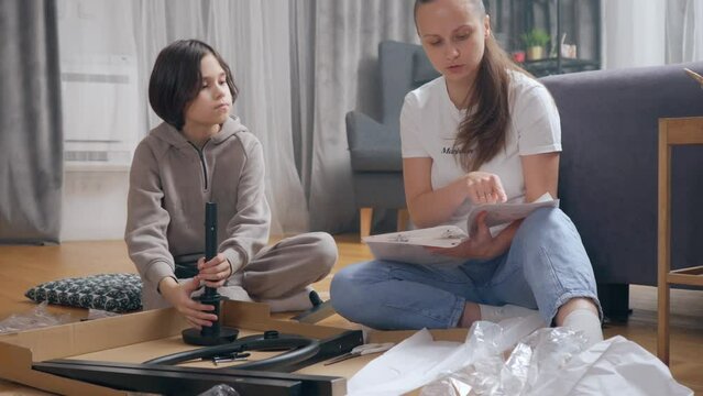 The mother explains and shows the child the correct order of assembly of parts, referring to the instructions for self-assembly of furniture, sitting with her son on the floor in the room.