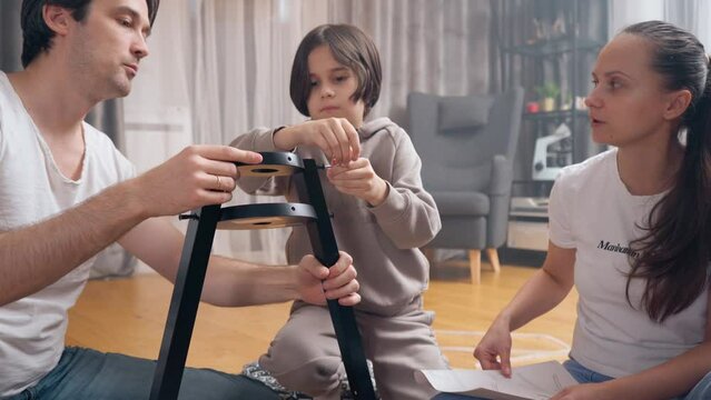 A little boy helps his parents assemble new furniture by screwing a screw. Parents spend time at home with their child, teaching him handmade skills in a playful way while sitting on the floor.