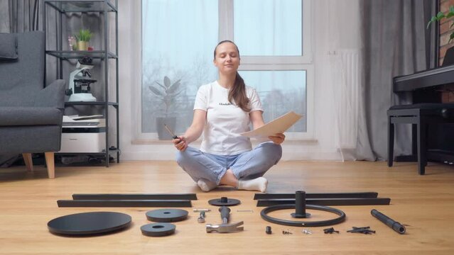A woman with her eyes closed meditates contentedly while sitting with instructions and a screwdriver in her hands in front of a set of parts for self-assembly of furniture at home.