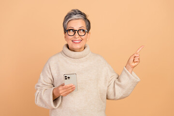 Portrait of old woman gray short hair wear sweater hold phone point finger interested media marketing isolated on beige color background