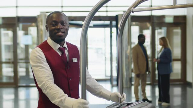 Portrait of young African American porter in uniform and white gloves standing with luggage cart in hotel and posing for camera with smile