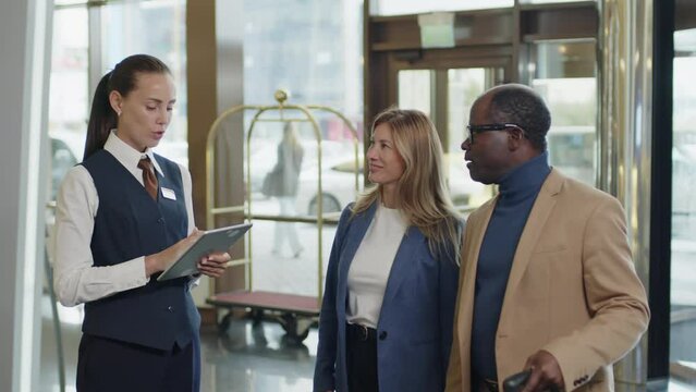 Female hotel manager standing in lobby, checking reservation number on digital tablet and welcoming arrived guests