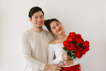Happy Asian couple holding red roses, both woman and man smiling and looking at camera, enjoy good time together on Valentines day, proposal and engage isolated over white background wall.