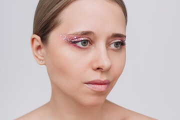 Young caucasian woman with purple and pink eye shadow and glitter on the eyelids on a gray...