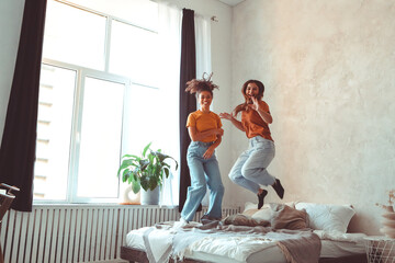Two happy joyful girlfriends of different races jumping on bed and having fun together at home - obrazy, fototapety, plakaty