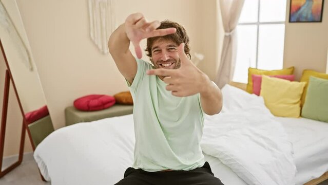 Cheery young caucasian man in pyjamas framing a photo concept with hands and fingers in bedroom, embracing his creativity with a bright smile