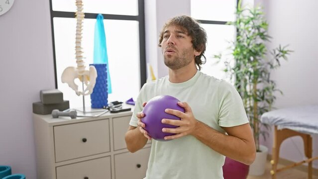 A young caucasian man with a beard holds a purple exercise ball in a rehab clinic room with medical equipment in the background.