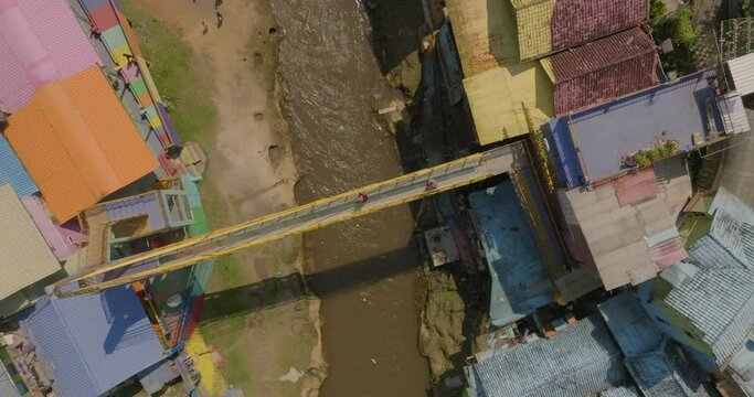 Aerial Top Panning Shot Of People Walking On Bridge Over Canal In Famous Kampung Pelangi Village, Drone Flying Upwards During Sunny Day - Java, Indonesia