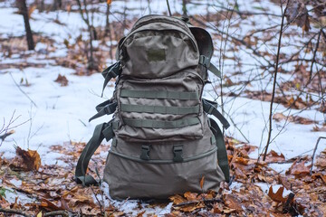 one large green army backpack stands on the ground in brown fallen leaves and white snow in the winter forest