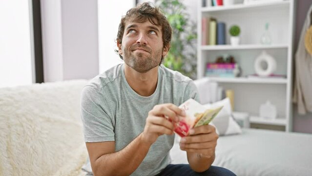 Young man, deep in thought, sits at home on sofa counting israeli shekel banknotes, contemplating his financial wealth and investment strategy
