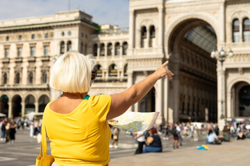 Senior Women in sunglasses looks at the paper map on Duomo square, Galleria Vittorio Emanuele II, Italy. Traveling Europe in summer. Blonde female is exploring new city and pointing finger