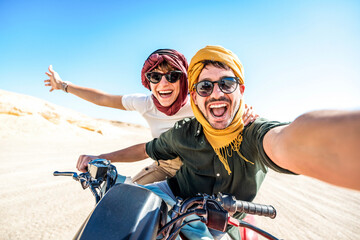 Young couple on a off road adventure excursion outside - Joyful tourists taking selfie with smart mobile phone in the desert - Tourism tour activities, transportation and summertime holidays concept - obrazy, fototapety, plakaty